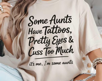 May include: Beige t-shirt with the text "Some Aunts Have Tattoos, Pretty Eyes & Cuss Too Much" in black script. Below the text is the phrase "it's me, I'm some aunts". The shirt is a casual style.