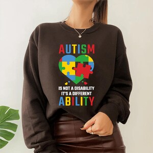 Peut inclure: Un sweat-shirt marron foncé avec le mot "AUTISME" en lettres colorées au-dessus d'un motif de puzzle en forme de cœur. Le texte "CE N'EST PAS UN HANDICAP, C'EST UNE CAPACITÉ DIFFÉRENTE" est en dessous.