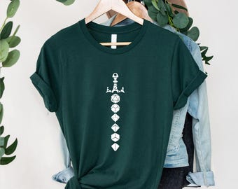 May include: A dark green t-shirt featuring a white graphic of a sword with dice. The design includes various dice shapes, perfect for tabletop gaming enthusiasts. The shirt is displayed on a wooden hanger.