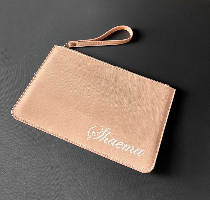 Op de afbeelding: Een lichtroze clutch met een polsbandje en de naam "Shaema" geborduurd in wit.