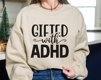 May include: A beige sweatshirt with the text "GIFTED with ADHD" in black. The sweatshirt is a crew neck style and is being worn by a person. The text is in a bold, sans-serif font.