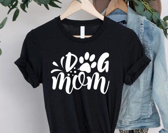 Może przedstawiać: Czarny t-shirt z białym napisem "Dog Mom" i grafiką łap. Koszulka wisi na drewnianym wieszaku. Rękawy są podwinięte.