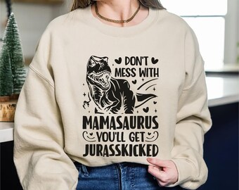 May include: A beige sweatshirt with a black graphic of a Tyrannosaurus Rex and the text "DON'T MESS WITH MAMASAURUS YOU'LL GET JURASSKICKED". The design includes hearts and a smiling face. The sweatshirt is worn by a person.
