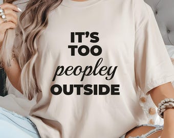 May include: A beige t-shirt with the phrase "IT'S TOO peopley OUTSIDE" printed in black. The shirt has short sleeves and a relaxed fit. The text is centered on the front of the shirt.