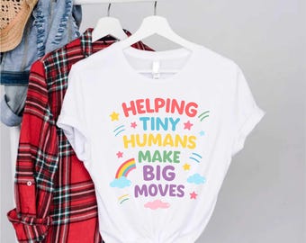 May include: White t-shirt with the text "Helping Tiny Humans Make Big Moves" in colorful letters. The design includes stars, a rainbow, and clouds. Other clothing items hang on a rack in the background.