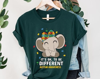 May include: A dark green short-sleeve t-shirt featuring a cartoon elephant with a colorful puzzle-piece patterned bow. The text reads IT'S OK, TO BE DIFFERENT AUTISM AWARENESS, surrounded by small scattered puzzle pieces in red, blue, and yellow.