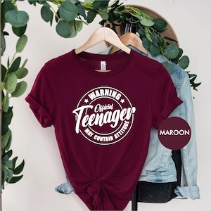 May include: A maroon t-shirt with a white circle graphic that says "Warning Official Teenager May Contain Attitude".