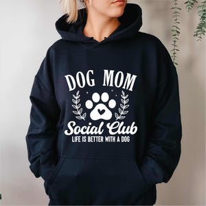 Puede incluir: Sudadera con capucha azul marino con el texto "DOG MOM Social Club" en blanco. El diseño incluye una huella de pata con un corazón, con las palabras "LIFE IS BETTER WITH A DOG". Hojas de laurel rodean el diseño. Prenda cómoda.