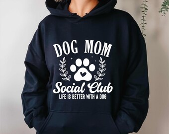 May include: A navy blue hooded sweatshirt with the text "DOG MOM Social Club" in white. A paw print with a heart is in the center of the design, with the words "LIFE IS BETTER WITH A DOG" below. The design is surrounded by laurel leaves.