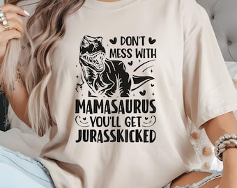 May include: A beige t-shirt with a black graphic of a Tyrannosaurus Rex and the text "Don't Mess With Mamasaurus You'll Get Jurasskicked". The design includes hearts and stars.
