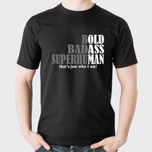 Può includere: T-shirt nera con le parole "BOLD BADASS SUPERHUMAN" impilate verticalmente in testo bianco e grigio. Sotto, la frase "that's just who I am!"