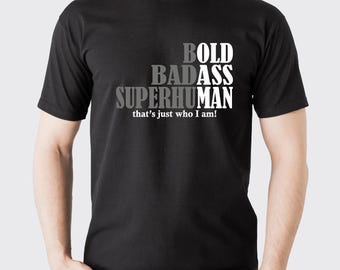 May include: Black t-shirt with the words "BOLD BADASS SUPERHUMAN" stacked vertically in white and gray text. Below the text is the phrase "that's just who I am!"