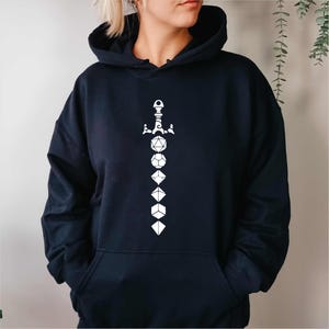 Puede incluir: Una sudadera con capucha azul marino con un gráfico blanco de una espada y dados. El diseño presenta una espada en la parte superior, con varias formas de dados que descienden por el centro. La sudadera tiene un bolsillo delantero y una capucha.