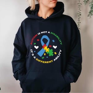 Puede incluir: Sudadera con capucha azul marino con un gráfico circular que dice "El autismo no es una discapacidad, es una capacidad diferente". El gráfico presenta una cinta azul, piezas de rompecabezas, corazones y estrellas. La sudadera tiene capucha y bolsillo delantero.