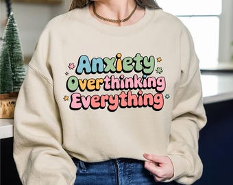 May include: A beige sweatshirt with the words "Anxiety Overthinking Everything" in colorful, retro-style lettering. The text is outlined in black and decorated with small star and flower graphics.