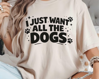 May include: Cream-colored t-shirt with the phrase "I JUST WANT ALL THE DOGS" in bold black letters. The design includes paw prints and stars. The shirt has short sleeves and a relaxed fit.