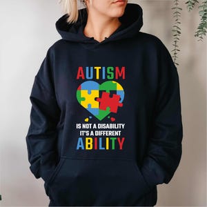 Puede incluir: Sudadera con capucha azul marino con la palabra "AUTISM" en rojo sobre un diseño de rompecabezas en forma de corazón en amarillo, verde, azul y rojo. El texto "IS NOT A DISABILITY IT'S A DIFFERENT ABILITY" está debajo.