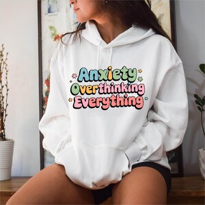 Puede incluir: Sudadera con capucha blanca con el texto "Anxiety Overthinking Everything" en letras de colores. La sudadera tiene un bolsillo delantero y una capucha con cordón. El texto está adornado con pequeñas estrellas y flores.