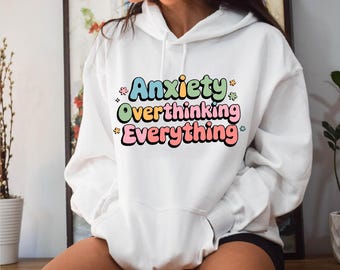 May include: White hoodie with the text "Anxiety Overthinking Everything" in colorful bubble letters. The hoodie has a front pocket and a drawstring hood. The text is surrounded by small star and flower graphics.