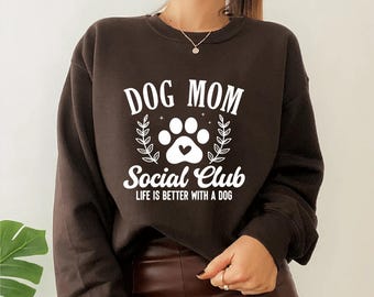 May include: A dark brown sweatshirt with the text "DOG MOM Social Club" in white, featuring a paw print and leaf design. The phrase "LIFE IS BETTER WITH A DOG" is printed below the club name. A casual, comfortable top.