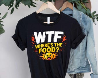 May include: Black t-shirt with the text "WTF WHERE'S THE FOOD?" in white, yellow, and orange. The design includes a grumpy face emoji. The shirt is hanging on a wooden hanger.