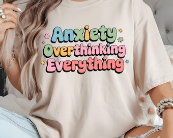 May include: Cream-colored t-shirt with the words "Anxiety Overthinking Everything" in colorful, retro-style lettering. The text is stacked in three lines, with each word in a different color and outlined in black. Small star and flower graphics are scattered around the text.