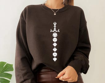 May include: A dark brown sweatshirt featuring a white graphic of a sword with dice. The dice include a D20, D12, D10, D8, D6, and D4. The sweatshirt is a crew neck style.