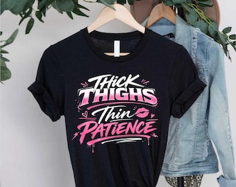 May include: Black t-shirt with the text "Thick Thighs Thin Patience" in white and pink lettering. The design includes a heart and a pair of lips. The shirt is hanging on a wooden hanger.