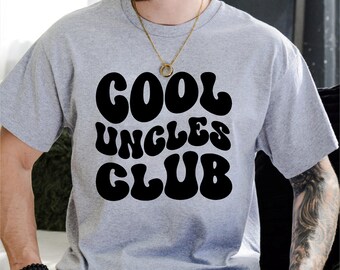 May include: A light gray t-shirt with the words "COOL UNCLES CLUB" in large, black, bubble letters. The shirt has short sleeves and a crew neck. The overall style is casual and comfortable.