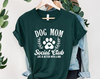 May include: A dark green t-shirt with the text "DOG MOM Social Club" in white. The design includes a paw print with a heart and the phrase "LIFE IS BETTER WITH A DOG". The shirt is hanging on a wooden hanger.