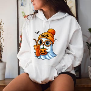 May include: White hoodie with a cartoon ghost design. The ghost wears an orange beanie with "BOO!" on it and glasses, holding a pumpkin and a drink. Two black bats fly above. The hoodie has a front pocket and is a casual garment.