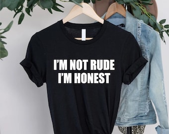 May include: Black t-shirt with the white text "I'M NOT RUDE I'M HONEST". The shirt is hanging on a wooden hanger. A denim jacket and a hat are visible in the background.