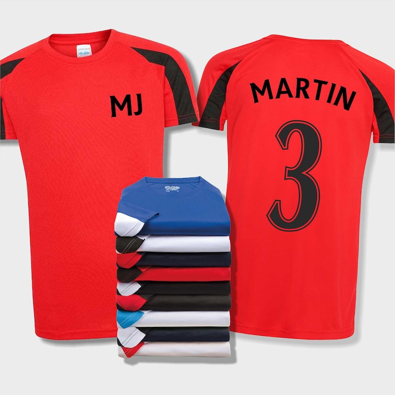 May include: Two red soccer jerseys with black and white accents. The front jersey has the initials "MJ" printed on the chest. The back jersey has the name "MARTIN" and the number "3" printed on the back. A stack of different colored jerseys is in the background.