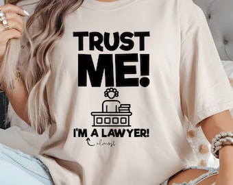 May include: A beige short-sleeved t-shirt featuring black text that reads TRUST ME! I'M A LAWYER! with the word almost written below with an arrow. A black graphic icon of a person sitting at a desk with books is positioned between the text lines.