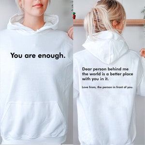 May include: White hoodie with the text "You are enough." on the front and the text "Dear person behind me the world is a better place with you in it. Love from, the person in front of you." on the back.