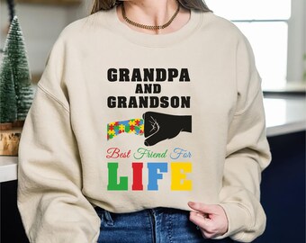 May include: A beige sweatshirt with the words "GRANDPA AND GRANDSON" in black, and "Best Friend For LIFE" in colorful letters. A black hand holds a puzzle piece design. The sweatshirt is worn by a person.
