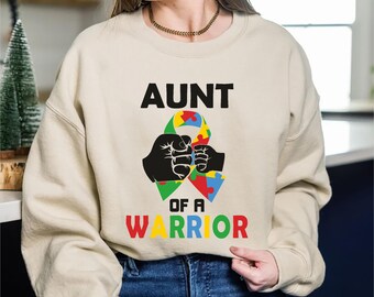 May include: Beige sweatshirt with the text "AUNT OF A WARRIOR" in black, red, yellow, green, and blue. The design includes two black fists holding a colorful puzzle ribbon, symbolizing autism awareness.