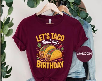 May include: Maroon t-shirt with the text "LET'S TACO bout my BIRTHDAY" in yellow. The graphic features two tacos with fillings, cacti, and chili peppers. The shirt is hanging on a wooden hanger.
