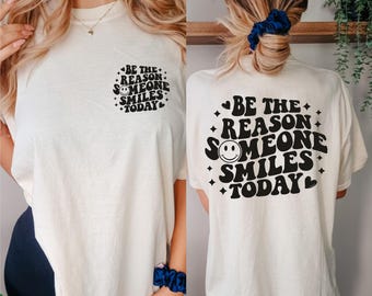 May include: Cream-colored t-shirt with the text "BE THE REASON SOMEONE SMILES TODAY" in black. The design includes a smiling face and decorative elements. The shirt is short-sleeved and has the same design on the front and back.