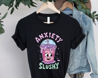 May include: A black short-sleeve t-shirt featuring a graphic of a pink slushy drink with a cartoon face, a striped straw, and the text "ANXIETY SLUSHY" in light purple and green lettering. The shirt is displayed on a wooden hanger.