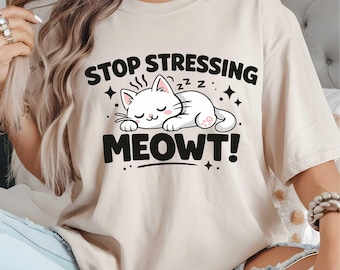 May include: A beige t-shirt with the text "STOP STRESSING MEOWT!" in black. The graphic features a sleeping white cat with pink accents, surrounded by stars. The shirt is a casual, comfortable style.