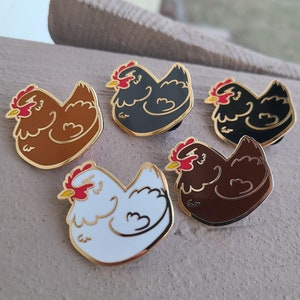May include: Five enamel pins featuring different colored chickens. The chickens are all in a sitting position with their heads tilted down. The colors of the chickens are brown, black, white, and two shades of brown.