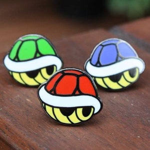 May include: Three enamel pins shaped like a turtle shell from the video game Super Mario Bros. The pins are green, red, and blue.