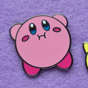 May include: Two enamel pins featuring characters from the video game Kirby. The first pin is pink and depicts Kirby with a surprised expression. The second pin is yellow and depicts a star-shaped character with a surprised expression.