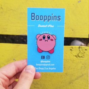 May include: Pink enamel pin of a cartoon character with large eyes and a small mouth. The character is pink with red accents. The pin is on a blue card with the text "Boopppins Enamel Pins" and contact information.