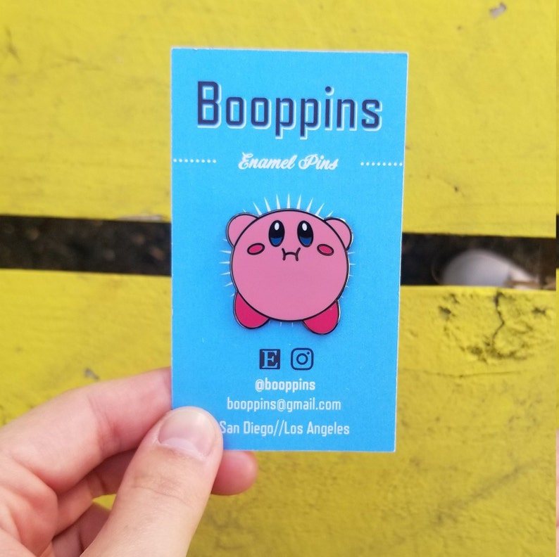 May include: Pink enamel pin of a cartoon character with large eyes and a small mouth. The character is pink with red accents. The pin is on a blue card with the text "Boopppins Enamel Pins" and contact information.