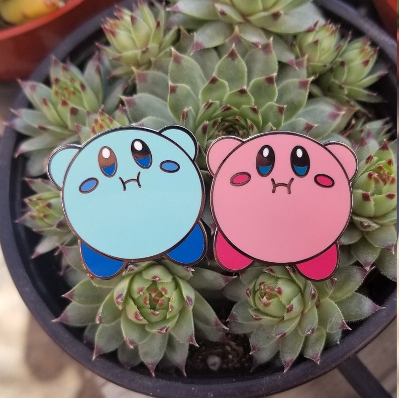 May include: Two enamel pins featuring the pink and blue characters from the video game Kirby. The pink character has a blush on its cheeks and both characters have a surprised expression.