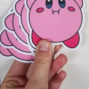 May include: A stack of pink and white stickers featuring a cartoon character with large eyes and a pink body. The character is smiling and has a small mouth.