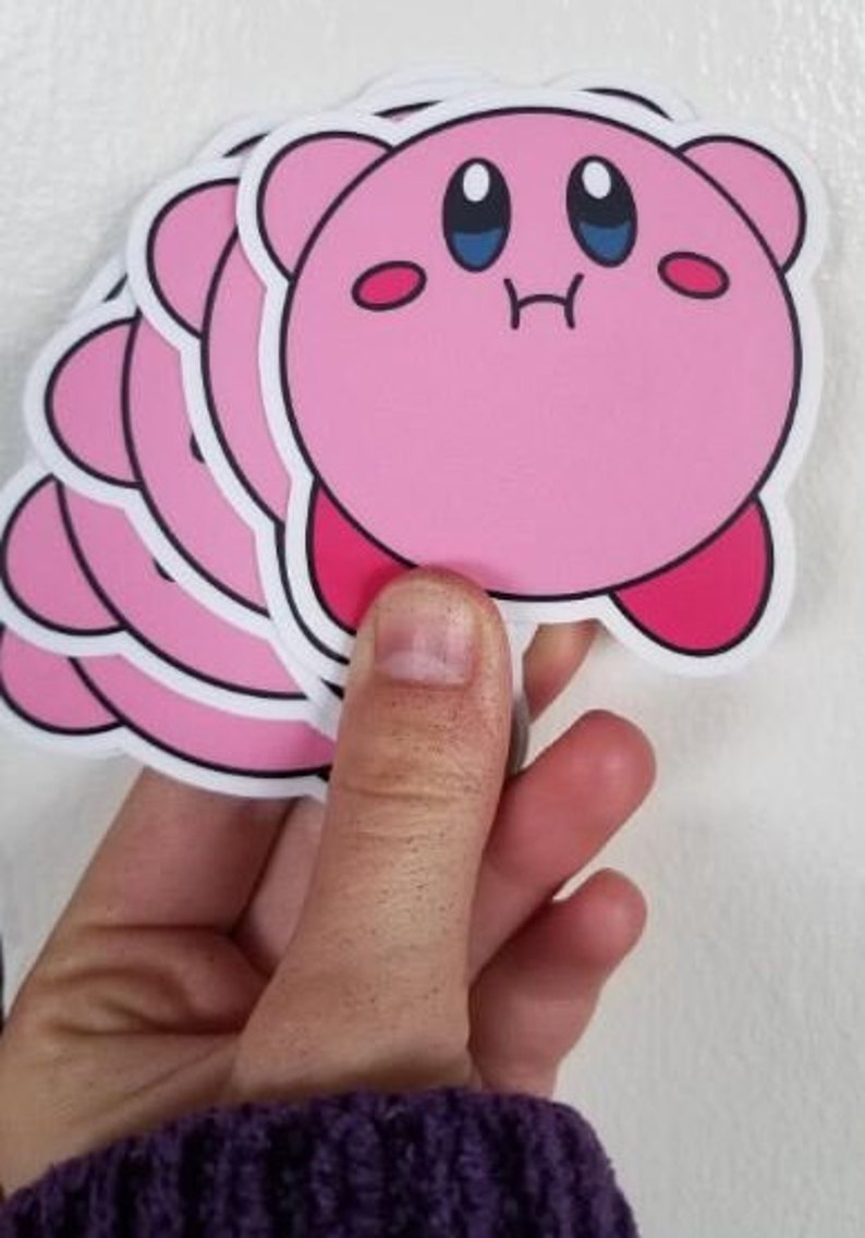May include: A stack of pink and white stickers featuring a cartoon character with large eyes and a pink body. The character is smiling and has a small mouth.