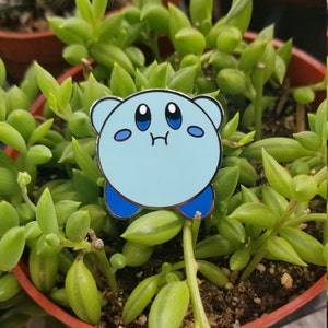 May include: A light blue enamel pin featuring a cartoon character with a sad expression. The character has large eyes, a small mouth, and a round body. The pin is set against a background of green succulent plants.
